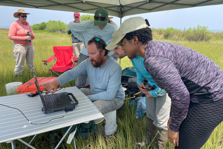 Several people huddle around a laptop computer that is open on a folding while out in the field. They are protected from the sun by a pop-up tent.