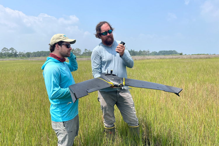 Two men stand in a marshy area. The man on the left is wearing a long-sleeved turquoise shirt and a tan baseball cap. The man on the right has on a long-sleeved light grey shirt. He is holding a large fixed-wing drone that looks like a miniature stealth airplane rather than the usual "quadcopter" drone