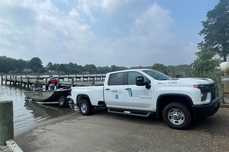 A white pickup truck backs a small boat into the water at a boat ramp. On the truck are logos from several organizations: The Virginia Institute of Marine Science, Chesapeake Bay National Estuarine Research Reserve Virginia, and NOAA.